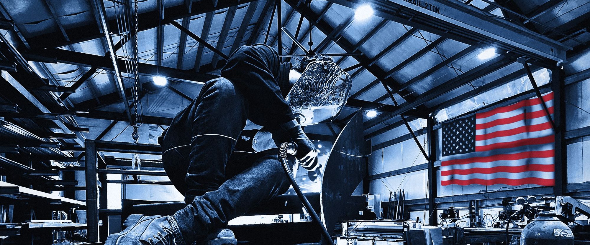 A welder in a hood works on a curved steel stair in the shop, with a United States flag on the far wall